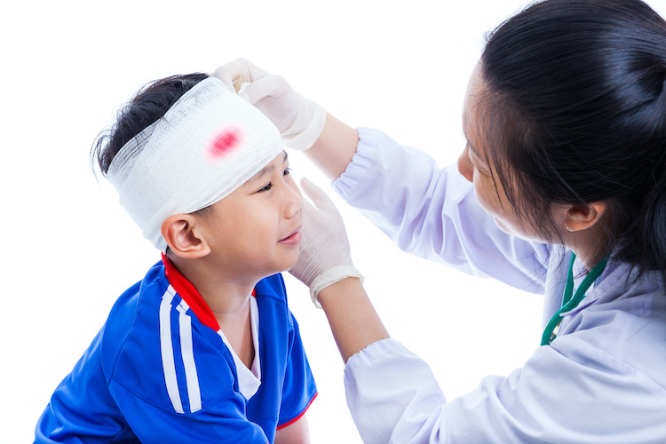 Sports injury. Athlete little asian (thai) boy in blue sportswear with trauma of the head crying, bloody on bandage. Doctor makes bandage on head patient, on white background. Studio shoot. Side view.